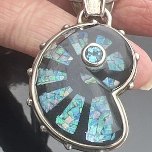 May include: A silver pendant featuring a black nautilus shell with a blue gemstone in the center. The shell is inlaid with iridescent opal pieces.