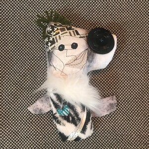 May include: A handmade plush toy with a white and black zebra print body, white fur trim, and a black button eye. The toy has a green and black patterned hat and a white face with embroidered details.