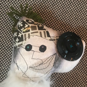 May include: A white fabric doll with a black and white patterned hat, a black button, and a stitched face with blue and purple eyes.