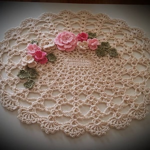 Può includere: Un centrino all'uncinetto con un delicato disegno floreale. Il centrino è di colore crema e presenta fiori all'uncinetto rosa e verdi.