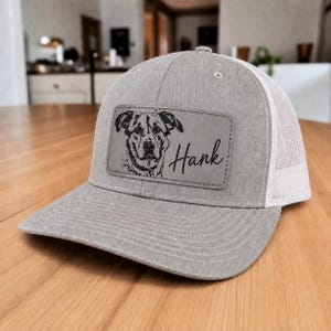 May include: A heather gray baseball cap with a white mesh back and a rectangular leather patch on the front. The patch features a black line drawing of a dog's face and the name Hank written in a cursive script font to the right of the dog.