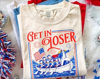 May include: An ivory crew neck t-shirt featuring a graphic print of George Washington standing in a boat with soldiers, holding an American flag. The text reads GET IN LOSER in red font. The design includes blue water, fireworks, and red borders.