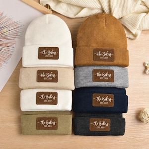 May include: A collection of eight folded knit beanies in various colors including white, tan, olive green, mustard yellow, gray, navy blue, and charcoal. Each beanie features a rectangular brown leather patch with the text the Bakers EST. 2019.