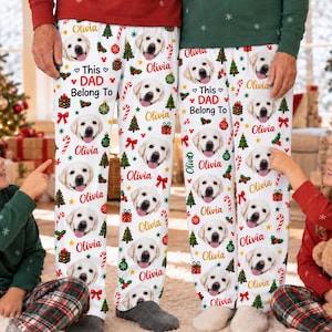 Pode incluir: Calças de pijama brancas com um padrão repetitivo de rostos de golden retriever, árvores de Natal, caixas de presente, bengalas doces e ornamentos, com o texto "This DAD Belong To" e o nome "Olivia" em vermelho e dourado.