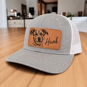 May include: A gray baseball cap with a white mesh back and a rectangular tan leather patch on the front. The patch features a black engraved illustration of a dog's face and the name Hank written in a cursive script font next to the dog image.