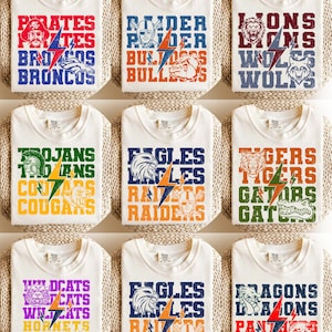 May include: Cream-colored t-shirts with various team names and mascots printed in bold, colorful fonts. Designs include "Pirates," "Broncos," "Bulldogs," "Lions," "Wolves," "Trojans," "Cougars," "Eagles," "Raiders," "Tigers," "Gators," "Wildcats," "Hornets," "Dragons," and "Panthers."