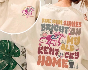 May include: A beige sweatshirt with a colorful graphic print on the back. The graphic features a horse, a jockey, and the text "The sun shines bright on my old Kentucky home".