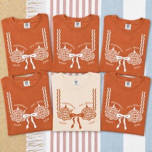 May include: Six crewneck t-shirts arranged in a grid. Five shirts are burnt orange with white graphic prints, and one center shirt is cream with burnt orange prints. Each features two disco ball pumpkins, a bow, and text reading Marianna's Fall Bach Salem 2026.