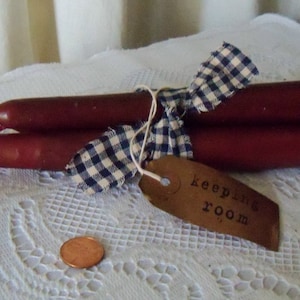 May include: Two burgundy taper candles tied together with a blue and white checkered ribbon. A brown tag with the words "keeping room" is attached. A penny is next to the candles.