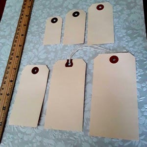 May include: A collection of six blank, rectangular price tags in varying sizes. Each tag is a light tan color with a reinforced brown eyelet. A wooden ruler with inch measurements is visible on the left side of the image.