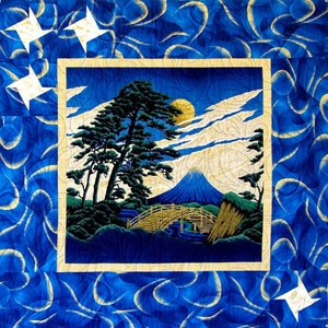 May include: A blue quilted fabric with a gold frame surrounding a picture of a mountain, trees, and a bridge. The picture is set against a blue sky with white clouds.
