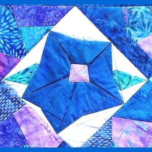 May include: A blue and purple patchwork quilt block with a geometric design. The block is made up of various shades of blue and purple fabric, with a white centre.