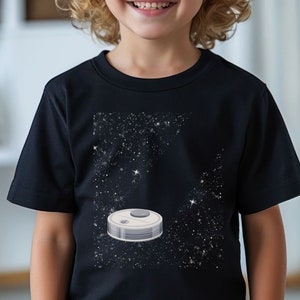 Peut inclure: Un t-shirt noir avec un motif spatial blanc et gris. Le motif représente un objet rond avec un petit cercle sur le dessus, entouré d'étoiles blanches sur un fond noir.