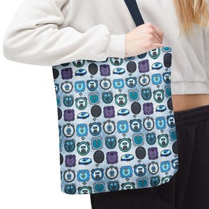 May include: A light blue tote bag with a repeating pattern of stylized faces in shades of blue, purple, and black. The bag has a dark blue shoulder strap and is held by a person.