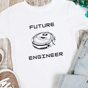 May include: White t-shirt with the text "FUTURE ENGINEER" and a graphic of a robot vacuum. The shirt is paired with light blue jeans and sparkly gold sneakers. The items are arranged on a light wood-grain surface.