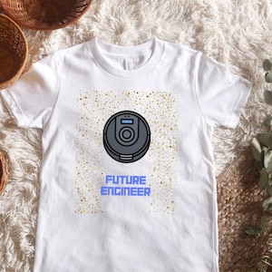 Puede incluir: Camiseta blanca con fondo de purpurina dorada y una ilustración en blanco y negro de un robot aspirador. El texto "FUTURE ENGINEER" está impreso en azul debajo del robot.