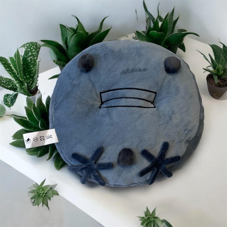 May include: A round, gray plush cushion with a cartoon face design. The cushion features dark blue accents for the eyes, mouth, and feet. The cushion is surrounded by green plants, creating a playful and inviting aesthetic.