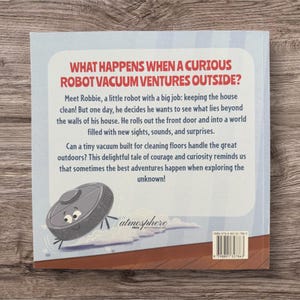 May include: Back cover of a children's book titled "What Happens When a Curious Robot Vacuum Ventures Outside?" The book features a cartoon robot vacuum illustration and text about adventure and curiosity. The book is on a wooden surface.