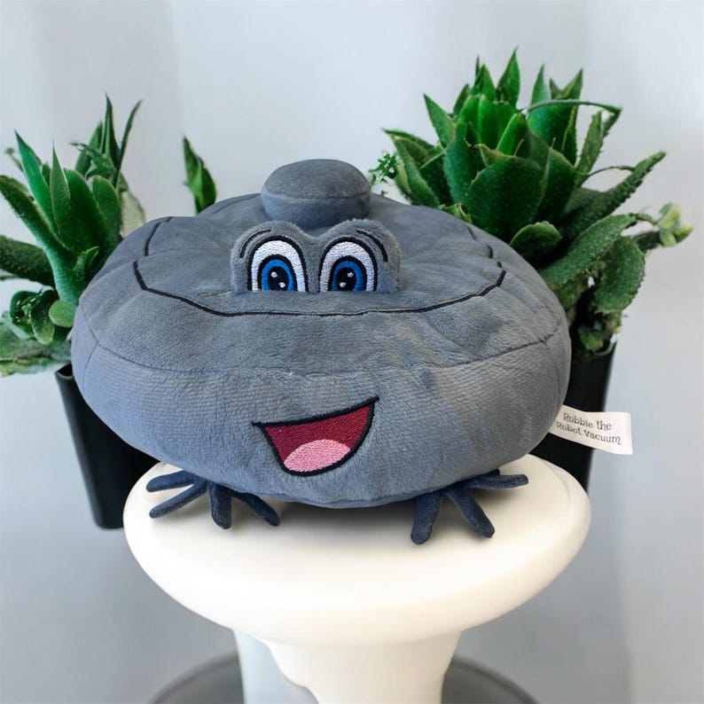 May include: A gray plush toy robot vacuum with a smiling face, blue eyes, and small black legs. The toy is sitting on a white surface, with green plants in black pots in the background. The tag reads "Robbie the Robot Vacuum".