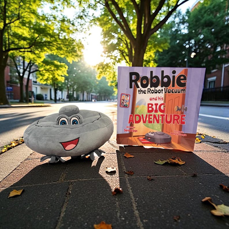 May include: A plush, gray robot vacuum toy with a smiling face and blue eyes sits next to a children's book titled "Robbie the Robot Vacuum and his BIG Adventure." The book cover features a cartoon illustration of a robot vacuum.