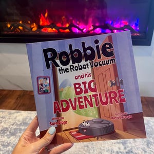 May include: A children's book titled "Robbie the Robot Vacuum and his Big Adventure." The cover features a cartoon robot vacuum, a red welcome mat, and the text "BIG ADVENTURE" in large letters. The book is written by Sara Biggs and illustrated by Daniela Frongia.