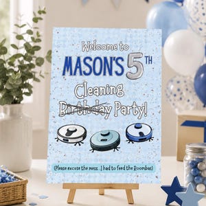 May include: A party sign on a small wooden easel, featuring the text "Welcome to MASON'S 5th Cleaning Birthday Party!" and illustrations of robot vacuum cleaners. The sign is light blue with blue and silver details. Balloons and decorations are in the background.