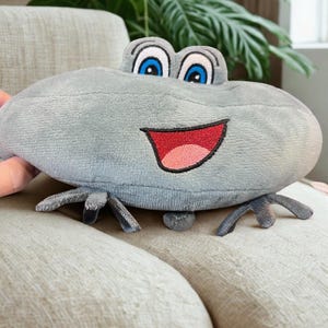 May include: A plush frog toy in shades of gray, featuring large blue eyes, a red smiling mouth, and small gray legs. The frog is sitting on a beige couch, with a green plant in the background.