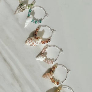May include: Five pairs of silver hoop earrings with different colored beads and seashells hanging from them. The beads are green, brown, and gold.
