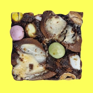 May include: A chocolate brownie topped with a variety of colourful candy eggs, including green, pink, and yellow. The brownie is topped with white chocolate and marshmallows.