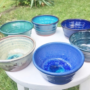 Pode incluir: Seis tigelas pequenas de cerâmica em vários tons de azul e verde. As tigelas estão dispostas em círculo sobre uma superfície branca.