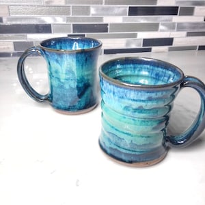 May include: Two handmade ceramic mugs with a blue and green glaze. The mugs have a rustic, handmade look and are perfect for coffee or tea.