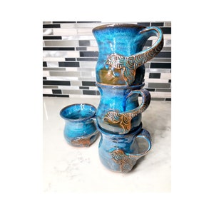 May include: Four handmade ceramic mugs and a small cup, stacked. The mugs are a striking blue with brown detailing and decorative handles. The small cup is next to the stack. The mugs are made of ceramic.