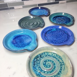 May include: A collection of six handmade ceramic spoon rests in various shades of blue and green. Each rest features a unique design, including swirls and textured patterns. The spoon rests are displayed on a white surface.