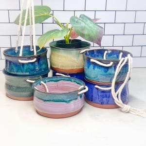 Op de afbeelding: Een verzameling handgemaakte keramische hangende plantenbakken in verschillende kleuren, waaronder blauw, groen en paars. Elke plantenbak heeft een uniek glazuur en wordt opgehangen met een wit touw. De plantenbakken zijn ontworpen voor planten.