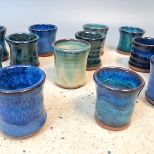 May include: A collection of small ceramic cups in various shades of blue and green. The cups are arranged in a semi-circle on a light-coloured surface.
