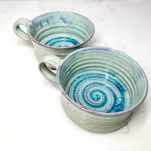 May include: Two handmade ceramic soup bowls with a light green glaze and a blue and purple swirl design. The bowls have a rounded shape and a handle.