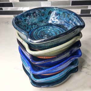 May include: A pile of handmade ceramic spoon rests in shades of blue and green. Each rest has a unique, glossy finish and a small handle. The rests are stacked, displaying the different colours and textures. The spoon rests are ideal for the kitchen.