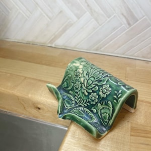 May include: A green ceramic dish with a floral design. The dish is shaped like a wave and has a raised edge. It is sitting on a wooden surface.