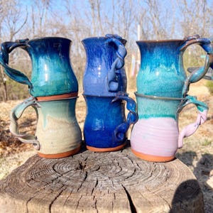 Puede incluir: Una pila de seis tazas de cerámica hechas a mano en varios tonos de azul, turquesa y rosa. Cada taza presenta un asa retorcida única y una base marrón en contraste. Las tazas se exhiben sobre una superficie de madera desgastada.