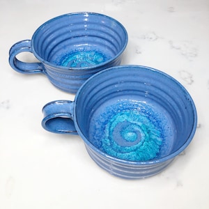 May include: A set of three ceramic mugs with a speckled, matte finish. The mugs are in shades of cream, light gray, and dark gray. Each mug has a simple, minimalist design and a rounded handle. The mugs are stacked on top of each other.