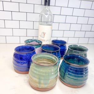 May include: A collection of handmade ceramic tumblers in various shades of blue, green, and purple, with a rustic, glazed finish. The tumblers are arranged around a wine bottle with a bird illustration on the label.
