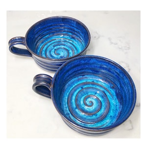 May include: Two handmade ceramic bowls with handles, featuring a vibrant blue glaze. The interior of each bowl showcases a spiral design in shades of blue and turquoise. The bowls are ideal for serving soups or stews.