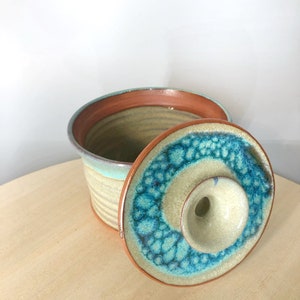 May include: A ceramic butter dish with a lid. The dish is glazed in a light blue and green colour with a brown rim. The lid has a small hole in the centre.