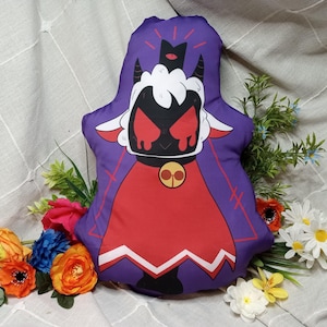May include: A plush pillow in the shape of a cartoon lamb with a black face, red eyes, and a red cape. The pillow is purple with a black crown and horns. The pillow is surrounded by colorful flowers.
