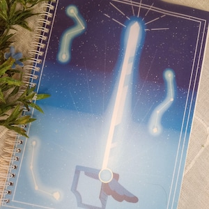 May include: A blue and white spiral bound notebook with a celestial design. The cover features a white sword with a blue handle and a starburst at the top. The background is a gradient of blue and purple with white stars.