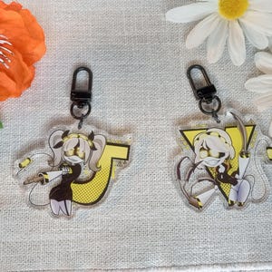 Peut inclure: Deux porte-clés en acrylique représentant un personnage de dessin animé en tenue noire et jaune. Les porte-clés ont un fermoir noir et sont décorés d'une lettre jaune. Des marguerites et une fleur orange sont en arrière-plan.