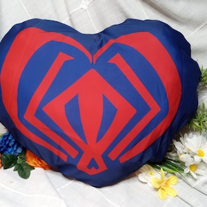 May include: Heart-shaped pillow in navy blue with a red geometric design. The pillow is surrounded by colorful flowers and greenery, creating a decorative and inviting aesthetic. The pillow is a soft furnishing item.