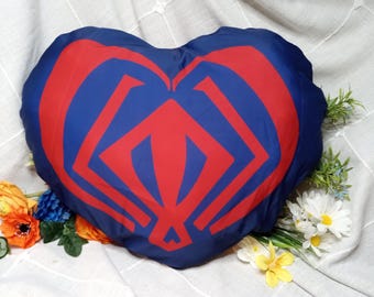 Puede incluir: Cojín en forma de corazón azul marino con un diseño geométrico rojo. El cojín está rodeado de flores y vegetación coloridas. Un artículo de decoración. Un cojín decorativo.