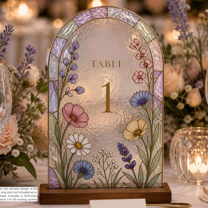 Peut inclure: Un panneau de table en verre en forme d'arche avec une bordure style vitrail et des illustrations florales colorées de marguerites, cosmos et lavande. Le centre affiche le texte doré "TABLE 1" et le panneau repose sur un socle en bois.