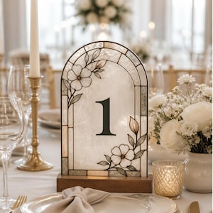 May include: A decorative stained glass table number featuring a floral motif and the number 1. The table number is set on a wooden base. Wedding decor includes candles, flowers, and glassware.